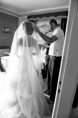 Photo en noir et blanc d’une mariée en robe de mariage avec un long voile, tandis qu’un homme ajuste le voile dans une chambre avant la cérémonie.