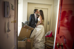 Un couple marié s'embrasse à l'intérieur d'un hall d'entrée d'immeuble rempli de cartons de déménagement et de sacs, avec un ascenseur en arrière-plan et une porte rouge brillante au premier plan.