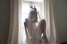Mariée assise sur une chaise près d’une fenêtre, enveloppée dans son voile, éclairée par la lumière naturelle.