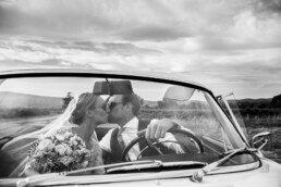 Mariés s’embrassant dans une voiture décapotable, la mariée tenant un bouquet, dans un paysage rural.