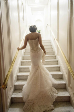 Mariée vue de dos montant un escalier en marbre, portant une robe de mariage en dentelle avec une longue traîne.