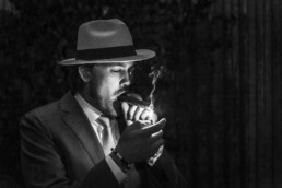 Portrait en noir et blanc d'un homme en costume et chapeau fedora allumant un cigare avec un briquet dans l'obscurité, la flamme éclairant son visage.