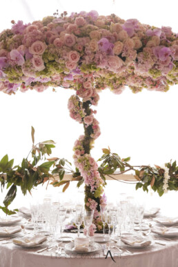 Centre de table de mariage spectaculaire en forme d'arbre fleuri composé de roses roses et d'orchidées, surmontant une table dressée avec de la vaisselle raffinée et des verres en cristal.