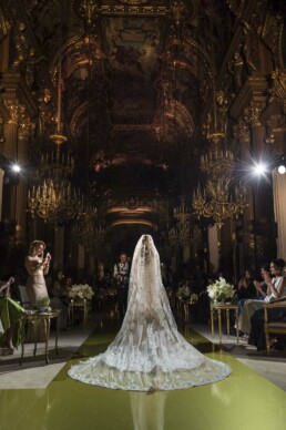 Vue de dos d'un mannequin portant une majestueuse robe de mariée au voile interminable brodé d'or, marchant sur un podium doré dans la salle de bal d'un opéra ou palais richement décoré.