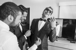 Un homme en costume de mariage élégant à motifs, portant des lunettes de soleil, rit aux éclats pendant que deux amis l'aident à s'habiller dans une chambre.