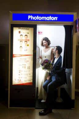 Un couple marié s'embrasse à l'intérieur d'un photomaton de marque 'Photomaton' avec des rideaux sombres. La mariée en robe de mariée blanche et le marié en costume s'embrassent, et un ami prend une photo de l'extérieur.