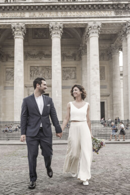 Un couple de mariés moderne marchant main dans la main sur les pavés devant les colonnes du Panthéon à Paris, la mariée portant un ensemble blanc élégant avec pantalon.