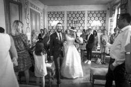 Le marié en costume sombre et la mariée en robe blanche tiennent un bouquet, entourés d'invités qui prennent des photos avec leurs téléphones.