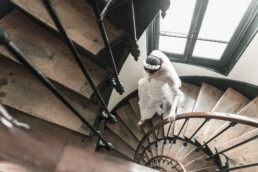 Mariée descendant un escalier en colimaçon en bois près d’une fenêtre lumineuse.