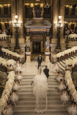 Mariée descendant un grand escalier fleuri entourée de petites danseuses en tutu dans une salle d’opéra.