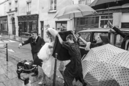 Mariée protégée de la pluie par ses proches avec des parapluies en sortant d’une voiture dans une rue parisienne.
