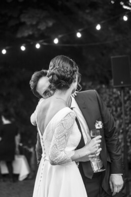 Mariée en robe de dentelle étreignant le marié lors d’une réception de mariage en extérieur, sous des guirlandes lumineuses.