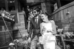 Couple de jeunes mariés souriants sortant d’un bâtiment entourés de leurs invités lors d’une célébration de mariage.