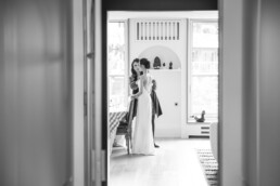 Une mariée en robe longue est aidée par une femme pour ajuster sa tenue dans une pièce lumineuse.