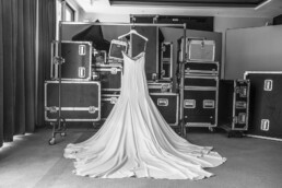 Robe de mariée suspendue devant des caisses techniques dans une pièce en noir et blanc.