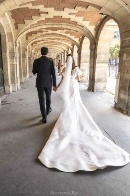 Un couple de mariés marche main dans la main sous une longue galerie d’arcades en pierre.
