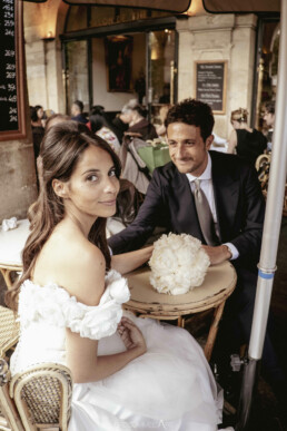 Un couple de mariés assis à la terrasse d’un café, la mariée regardant l’objectif tandis que le marié lui sourit.
