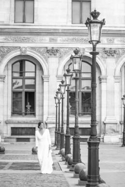 Une mariée en robe blanche marche sur une place pavée bordée de lampadaires devant un bâtiment classique.
