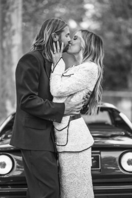 Un couple de mariés s’embrasse devant une voiture.