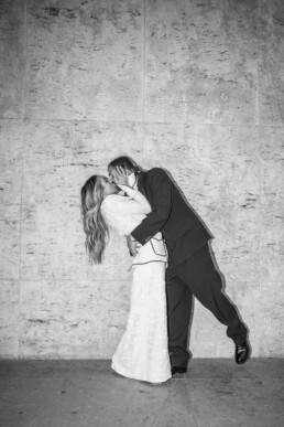 Un couple de mariés s’embrasse avec énergie devant un mur en pierre clair.